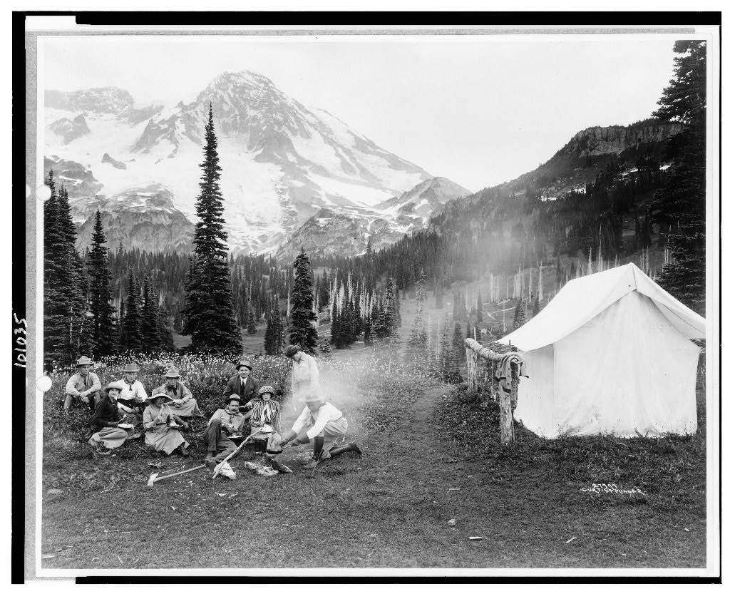 Camping party of men and women cooking at campfire and eating near tent in Indian Henry, Mt. Rainier National Park, Washington